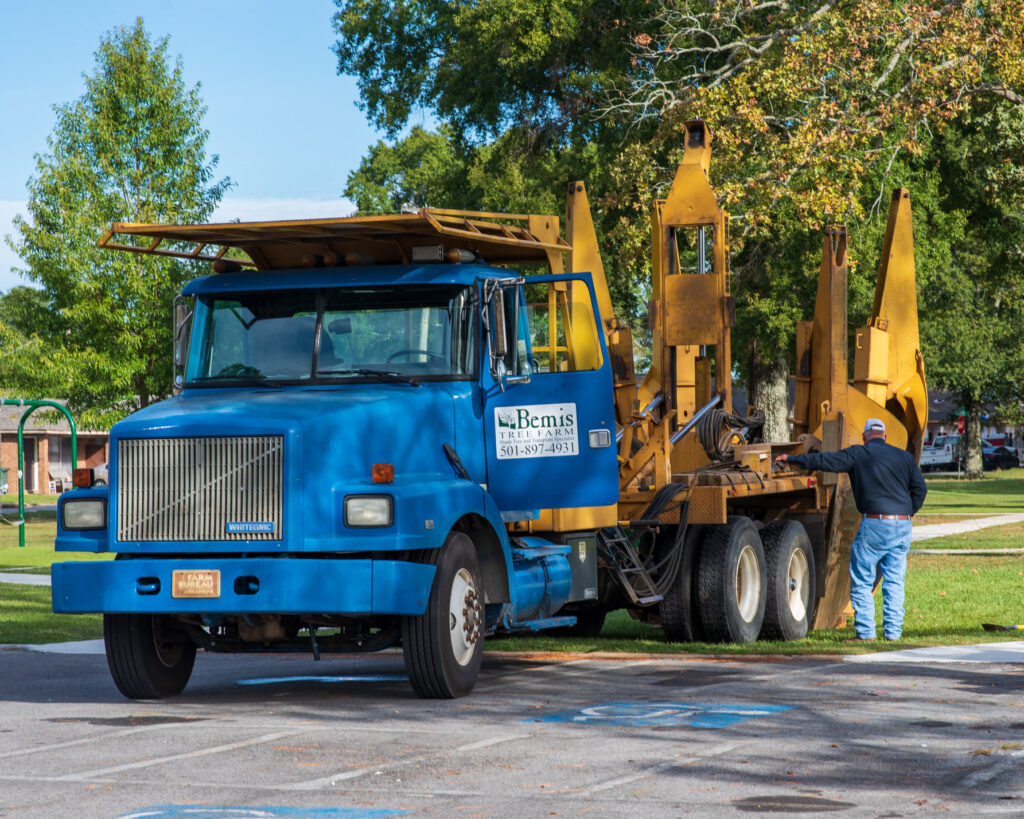 A man operates a large, blue tree transplanting truck in a park, with equipment raised and trees in the background.